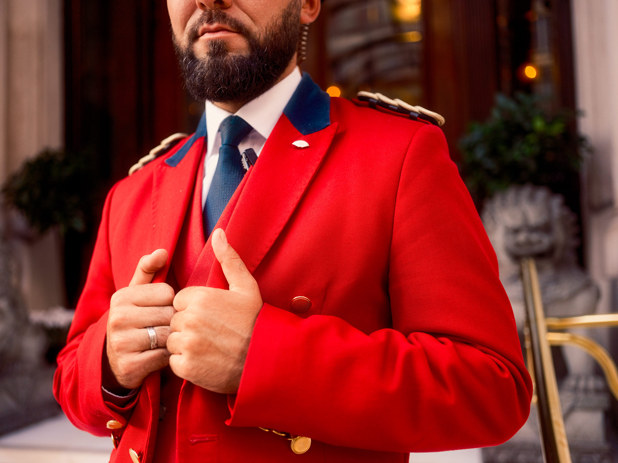 A doorman wearing a bright red jacket and resting his hands in pockets.